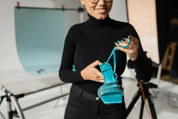 Обрезанный вид улыбающегося африканского производителя контента в черной водолазке с модной сандалией в размытой фотостудии — стоковое фото