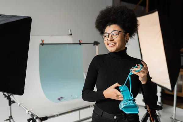 Happy african american content maker in eyeglasses holding stylish shoe and looking away near reflectors and shooting table in photo studio — Stock Photo