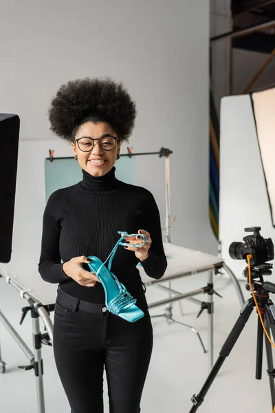 Satisfeito Africano americano gerente de conteúdo em óculos segurando sapato elegante e sorrindo para a câmera no estúdio de fotos moderno — Fotografia de Stock