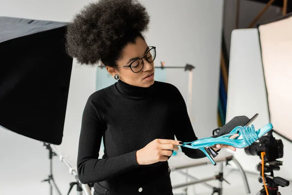 Fabricante de contenido afroamericano en gafas y cuello alto negro mirando calzado elegante en estudio de fotos borrosas - foto de stock