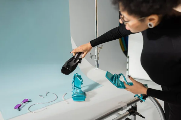 Fabricante de conteúdo afro-americano desfocado segurando calçados na moda perto de óculos de sol e acessórios de jóias na mesa de tiro no estúdio de fotos — Fotografia de Stock