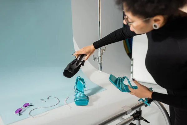 Fabricant de contenu afro-américain flou tenant des chaussures à la mode près de lunettes de soleil et accessoires de bijoux sur la table de tir en studio photo — Photo de stock