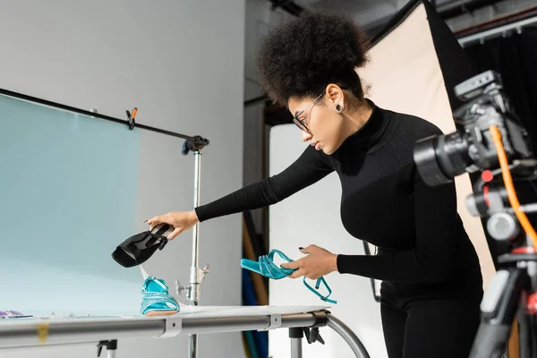 Producteur de contenu afro-américain tenant des chaussures à la mode près de la table de tir et appareil photo numérique flou dans le studio photo — Photo de stock