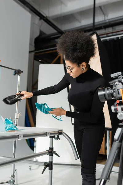 Gestionnaire de contenu afro-américain tenant sandales à la mode près de la table de tir et appareil photo numérique flou dans le studio photo — Photo de stock
