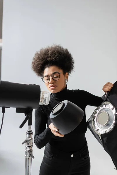 Gerente de contenido afroamericano en gafas con reflector mientras monta una lámpara estroboscópica en un estudio fotográfico - foto de stock