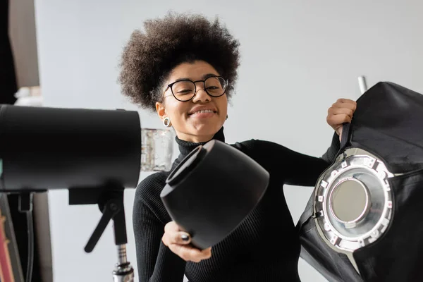 Carefree african american content producer holding softbox near strobe lamp and smiling at camera in photo studio — Stock Photo