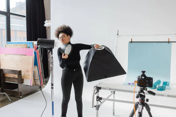 Produttore di contenuti afroamericani assemblaggio di apparecchiature di illuminazione e tenendo riflettore in studio moderno — Foto stock
