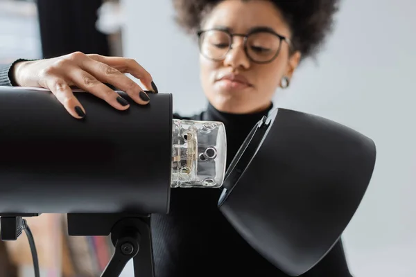 Productor de contenido afroamericano borroso en gafas que ensamblan la lámpara estroboscópica en el estudio fotográfico - foto de stock