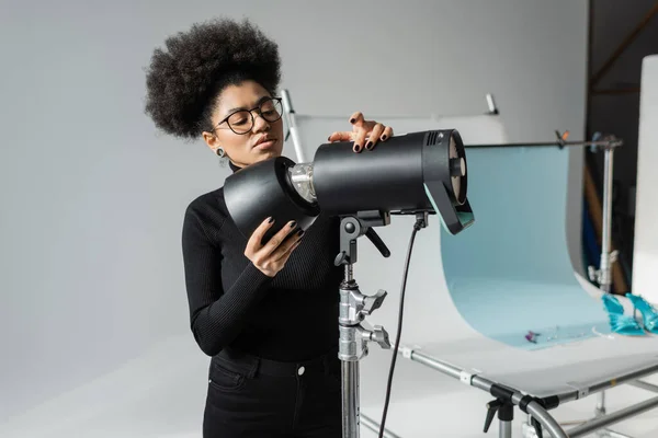 Gerente de conteúdo americano africano em óculos e gola alta preta trabalhando com lâmpada estroboscópica perto da mesa de tiro no estúdio de fotos — Fotografia de Stock