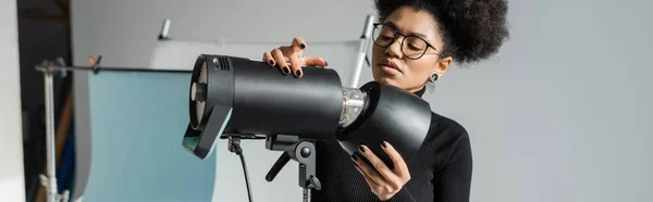 Fabricante de conteúdo americano africano elegante em óculos que trabalham com equipamentos de iluminação profissional no estúdio de fotografia, banner — Fotografia de Stock