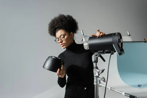 African american content maker in black turtleneck and eyeglasses holding detail of strobe lamp in photo studio — Stock Photo
