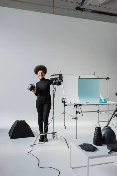 Pleine longueur de heureux fabricant de contenu afro-américain près de lampe stroboscopique et table de tir dans le studio photo contemporain — Photo de stock