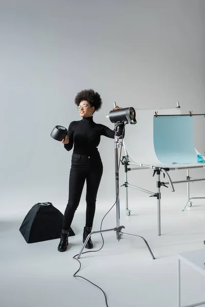 Full length of cheerful african american content producer assembling lighting equipment near shooting table in photo studio — Stock Photo
