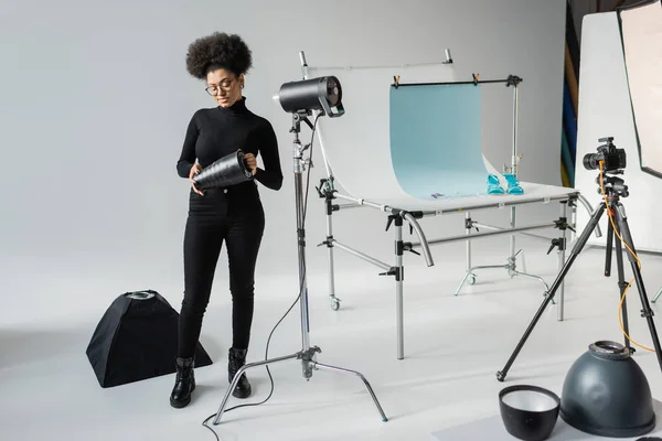 Intera lunghezza del produttore di contenuti afro-americani che detiene parte della lampada stroboscopica mentre lavora con apparecchiature di illuminazione in uno studio moderno — Foto stock