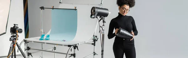 African american content manager assembling strobe lamp near digital camera and shooting table in photo studio, banner — Stock Photo
