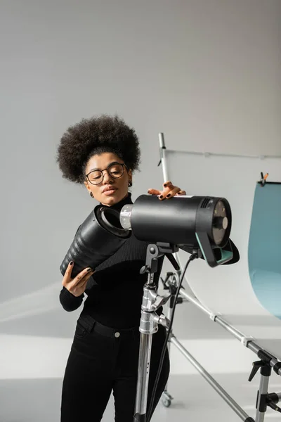 Productor de contenido afroamericano en gafas que montan foco estroboscópico en estudio fotográfico - foto de stock