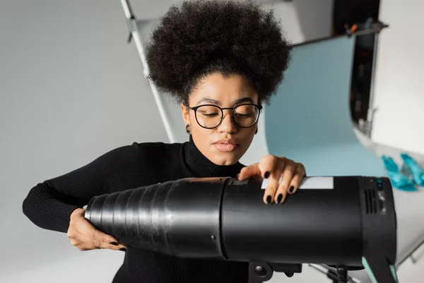 African american content producer in black turtleneck and eyeglasses assembling strobe spotlight in photo studio — Stock Photo
