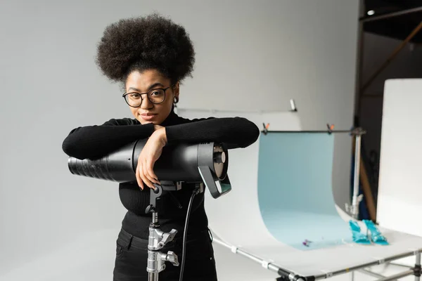 Elegante gerenciador de conteúdo afro-americano sorrindo para a câmera perto da lâmpada estroboscópica e mesa de tiro borrada no estúdio de fotos — Fotografia de Stock