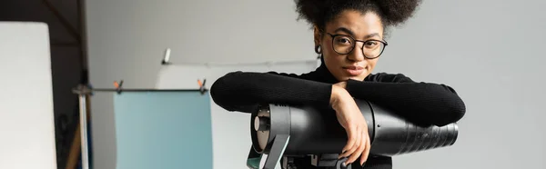 Positive african american content producer in stylish eyeglasses looking at camera near strobe lamp in photo studio, banner — Stock Photo