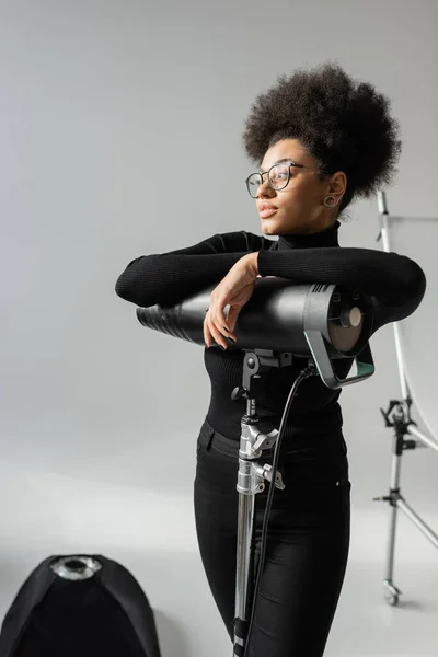African american content producer in eyeglasses and black turtleneck looking away near strobe floodlight in photo studio — Stock Photo