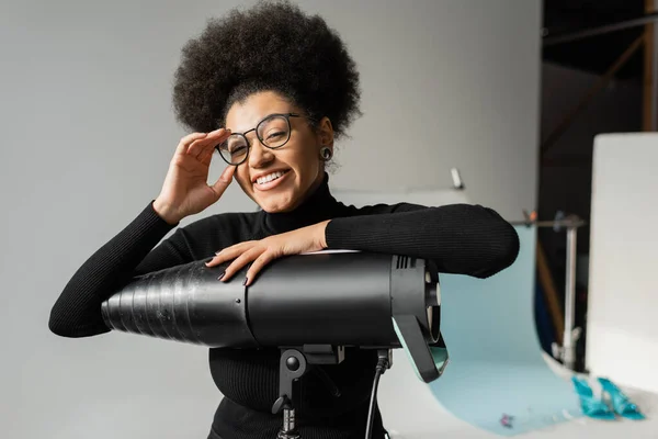 Fabricante de contenido afroamericano alegre ajuste de gafas con estilo y mirando a la cámara cerca de la lámpara estroboscópica en el estudio de fotos - foto de stock