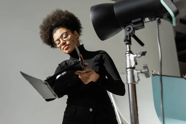 Producteur de contenu afro-américain dans les lunettes tenant une partie de lampe stroboscopique dans le studio photo — Photo de stock