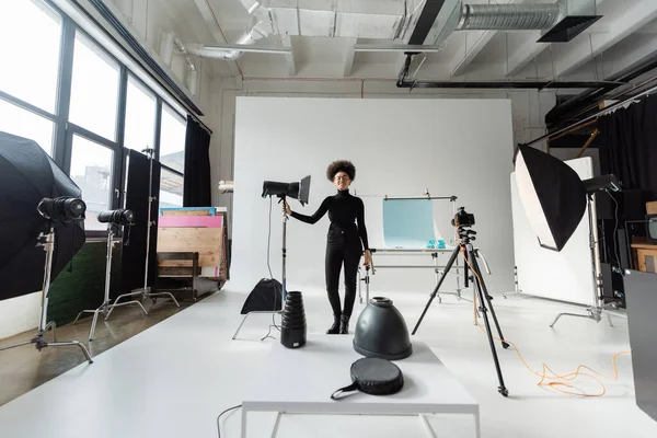 Piena lunghezza del felice gestore di contenuti africani americani in abiti neri in piedi vicino alla lampada stroboscopica in studio moderno — Foto stock
