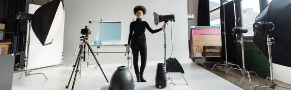 Longitud completa del gerente de contenido afroamericano feliz en ropa negra cerca del centro de atención y la cámara digital en el estudio de fotos moderno, pancarta - foto de stock