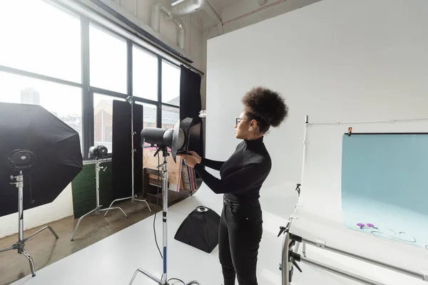Vista laterale del produttore di contenuti afro-americani che regola la lampada stroboscopica mentre lavora in uno studio fotografico contemporaneo — Foto stock