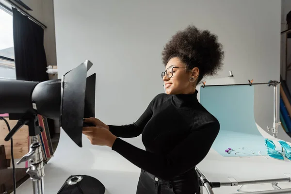 Fröhlich afrikanisch-amerikanischer Content-Produzent mit Sonnenbrille und schwarzem Rollkragen arbeitet mit Blitzlicht im Fotostudio — Stockfoto