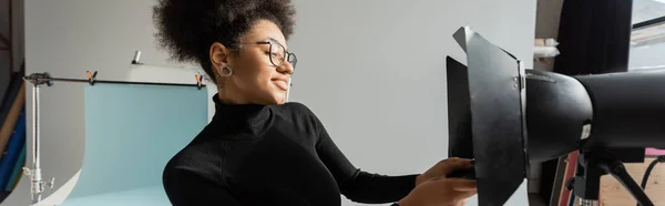 Pleased african american content maker in black turtleneck and eyeglasses working with strobe floodlight in photo studio, banner — Stock Photo