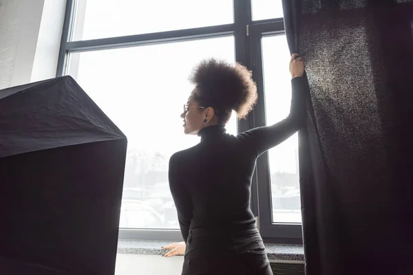 Rückansicht afrikanisch-amerikanischer Content-Produzent zieht Vorhang in Fensternähe bei Softbox-Reflektor im Fotostudio — Stockfoto