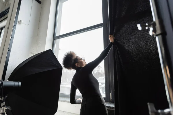 African American content manager in dolcevita nero tirando tenda vicino alla finestra vicino softbox riflettore in studio fotografico — Foto stock