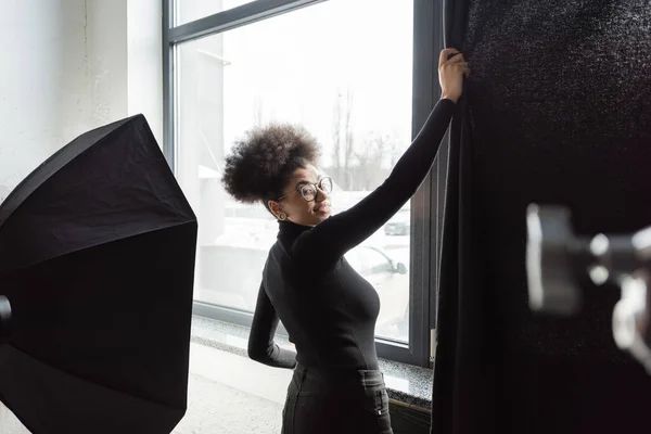 Produtor de conteúdo americano africano alegre puxando cortina perto da janela no estúdio de fotos e sorrindo para a câmera — Fotografia de Stock