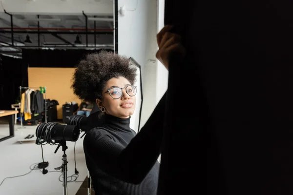 Fabricante de contenido afroamericano en gafas sonriendo cerca de cortina negra en estudio de fotos - foto de stock