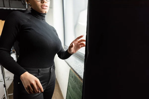 Vista recortada del fabricante de contenido afroamericano en cuello alto cerca de la ventana y cortina negra en el estudio de fotos — Stock Photo