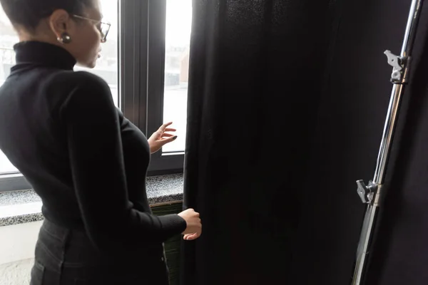 Афроамериканський менеджер контенту в турболенку, що стоїть біля вікна і чорної завіси в фотостудії — стокове фото