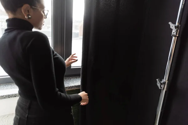 African american content manager in turtleneck standing near window and black curtain in photo studio — Stock Photo