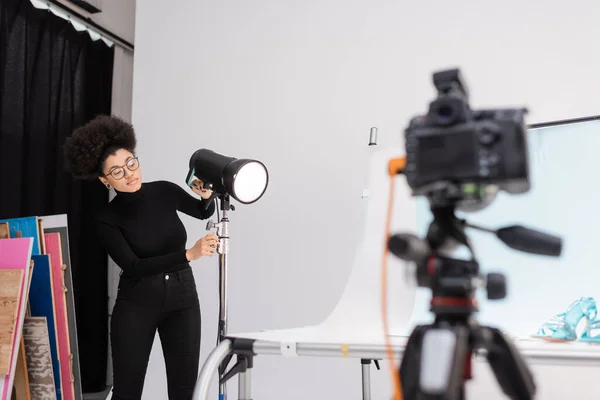 Afro-américain fabricant de contenu réglage projecteur près de la table de tir et appareil photo numérique flou dans le studio photo — Photo de stock