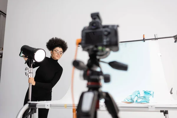 Afrikanisch-amerikanischer Content Manager justiert Blitzlampe in der Nähe verschwommener Digitalkamera und Aufnahmetisch mit stylischem Schuhwerk im Fotostudio — Stockfoto