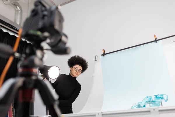 Afrikanisch-amerikanischer Content-Hersteller blickt auf verschwommene Digitalkamera in der Nähe von Scheinwerfern und Aufnahmetisch mit trendigen Schuhen im Fotostudio — Stockfoto