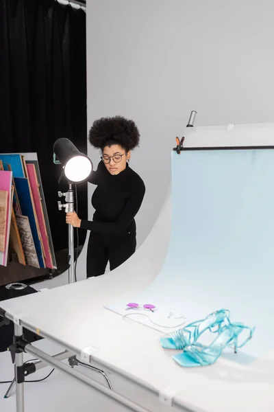 Produtor de conteúdo afro-americano em óculos perto de holofotes e óculos de sol elegantes com sandálias na mesa de tiro no estúdio de fotos — Fotografia de Stock