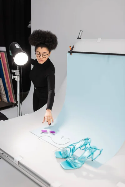 Productor de contenido afroamericano cerca de la lámpara estroboscópica y la mesa de tiro con gafas de sol y sandalias con estilo en el estudio de fotos - foto de stock