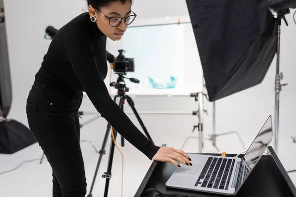 African american content producer in eyeglasses using laptop near reflector and blurred digital camera in photo studio — Stock Photo