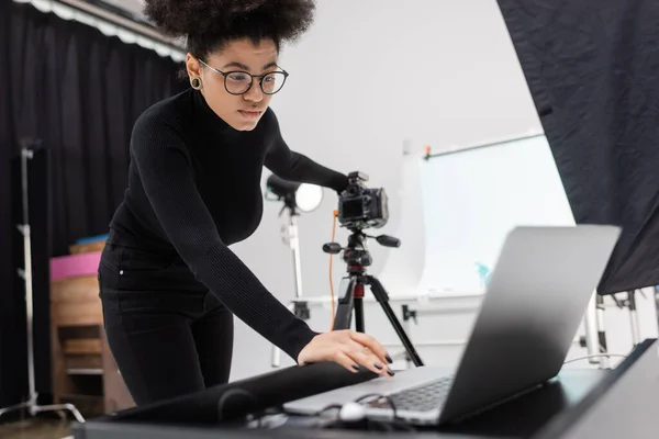 Produtor de conteúdo afro-americano em gola alta preta e óculos olhando para laptop enquanto trabalhava no estúdio de fotografia — Fotografia de Stock