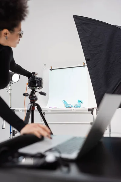 Афроамериканський виробник контенту, що налаштовує цифрову камеру біля зйомки столу з модним взуттям та розмитим ноутбуком у фотостудії — стокове фото