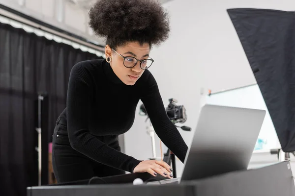 Producteur de contenu afro-américain concentré à l'aide d'un ordinateur portable dans un studio photo au premier plan flou — Photo de stock