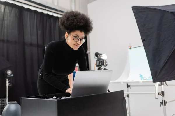 Sonriente afroamericano gerente de contenido en gafas y cuello alto negro mirando a la cámara cerca de la computadora portátil en el estudio de fotos - foto de stock