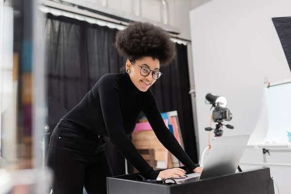 Fabricante de contenido afroamericano elegante y alegre mirando a la cámara y el uso de la computadora portátil en el estudio de fotos — Stock Photo