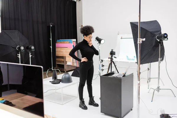 Full length of thoughtful african american content maker in black clothes standing with hand on hip near laptop in modern photo studio — Stock Photo